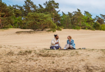 Dos personas disfrutan de un picnic sobre la arena junto a árboles en un alojamiento glamping.