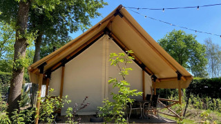 Safari-Glampingzelt mit Holzterrasse und Stühlen, umgeben von Bäumen, unter blauem Himmel.
