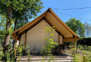 Tienda de glamping tipo safari con terraza de madera y sillas, rodeada de vegetación y luces.