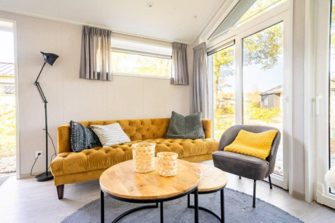 Salon lumineux et cosy du lodge Berkel à De Wije Werelt aux Pays-Bas, avec canapé moutarde et grandes fenêtres.