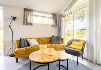 Salon lumineux et cosy du lodge Berkel à De Wije Werelt aux Pays-Bas, avec canapé moutarde et grandes fenêtres.