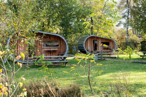 Dos pequeñas casas ovaladas de madera llamadas Boet en De Wije Werelt, Países Bajos, rodeadas de naturaleza verde.
