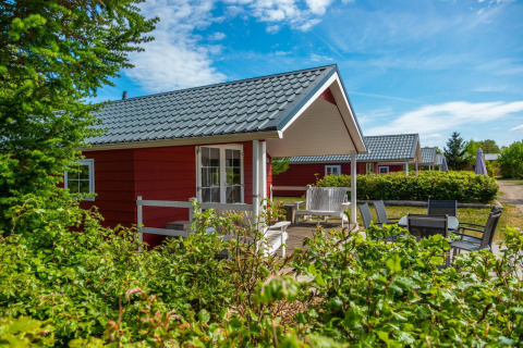 Charming red cabin at Boslodge with patio, outdoor seating, greenery, and a bright blue sky overhead.