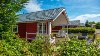 Acogedora cabaña roja en Boslodge con terraza, sillas de jardín y abundante vegetación bajo cielo azul.
