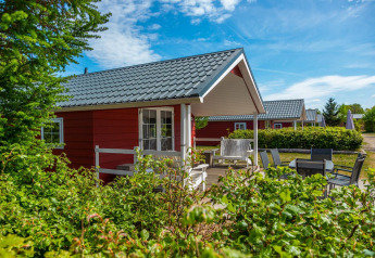 Knusse rode houten Boslodge met veranda, tuinmeubilair en veel groen onder een stralend blauwe lucht.