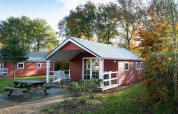 Rød Boslodge hytte ved De Wije Werelt i Holland, omgivet af træer med bordbænk og haveområde.