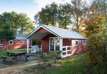 Rote Boslodge im De Wije Werelt, Niederlande, umgeben von Bäumen und mit einer Bank im Vordergrund.