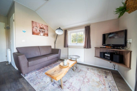 Soggiorno nella Boslodge a De Wije Werelt, Paesi Bassi, con divano, tavolo in legno e TV sul muro.