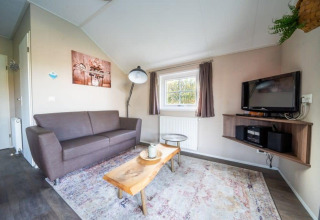 Soggiorno nella Boslodge a De Wije Werelt, Paesi Bassi, con divano, tavolo in legno e TV sul muro.