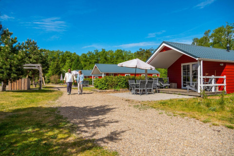 Dos personas caminan por un sendero frente a una cabaña roja Boslodge con terraza y muebles de jardín.