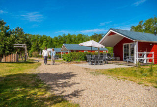 Two people walk on a path in front of a red Boslodge cabin with patio furniture, surrounded by greenery.