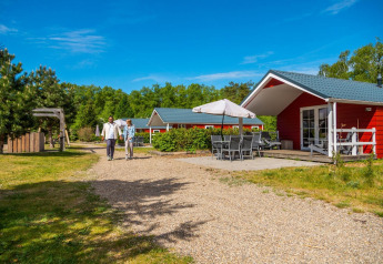 To personer går på en sti foran en rød Boslodge-hytte med terrasse og udendørs møbler omgivet af natur.