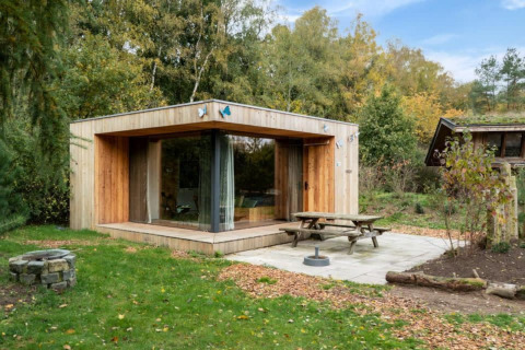 Cabaña moderna de madera llamada Fred en un entorno forestal, con grandes ventanales y mesa exterior.