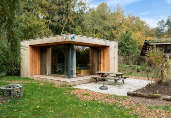 Moderne træhytte ved navn Fred i skovområde, med store vinduer, picnicbord og bålplads udenfor.