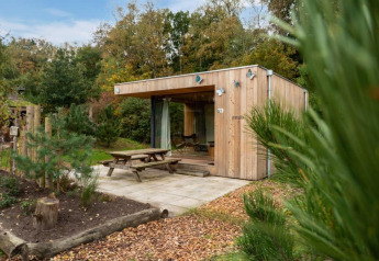 Trælodge Fred ved De Wije Werelt i Holland, omgivet af træer med udendørs picnicbord på terrassen.