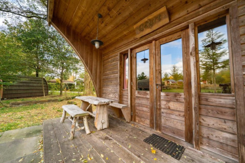 Porche de madera y banco frente a la tiny house Geurt en De Wije Werelt, Países Bajos, rodeada de naturaleza.