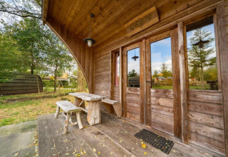 Porche de madera y banco frente a la tiny house Geurt en De Wije Werelt, Países Bajos, rodeada de naturaleza.
