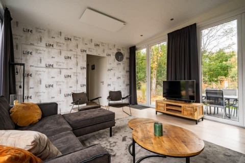 Wohnzimmer in der Lodge Just Nature in De Wije Werelt, Niederlande mit Sofa, TV, großen Fenstern und Holztisch.