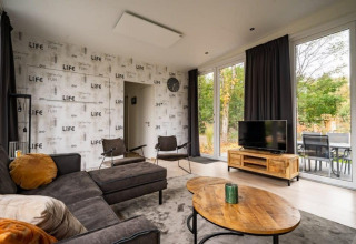 Woonkamer van lodge Just Nature bij De Wije Werelt, Nederland met bank, tv, grote ramen en houten meubilair.