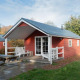 Boslodge, en rød træhytte med overdækket terrasse og bænk, omgivet af grønt og træer i naturen.