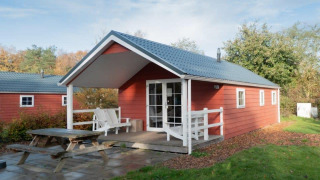 Boslodge, en rød træhytte med overdækket terrasse og bænk, omgivet af grønt og træer i naturen.