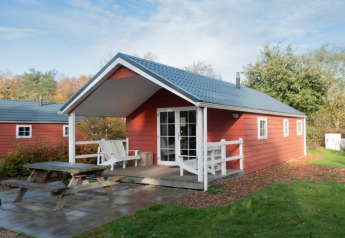 Boslodge, een rood houten chalet met overdekte veranda en picknickbank, omgeven door groen.