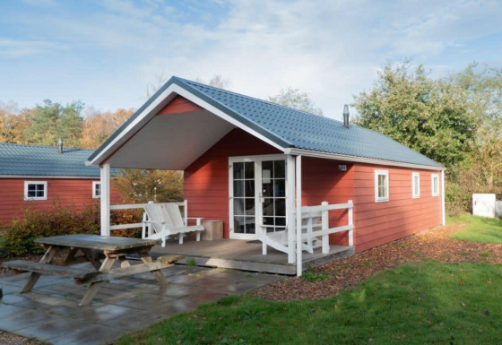 Boslodge, en rød træhytte med overdækket terrasse og bænk, omgivet af grønt og træer i naturen.