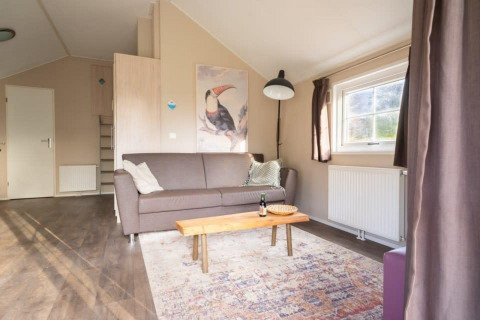 Sala de estar en Boslodge, De Wije Werelt, Países Bajos, con sofá, arte de tucán, mesa y ventanal.