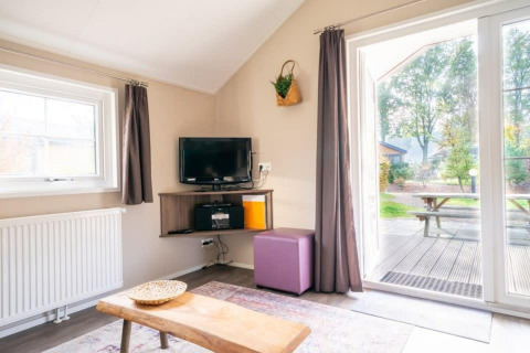 Interior luminoso de una Boslodge en De Wije Werelt, Países Bajos, con TV, mesa y vista al jardín.