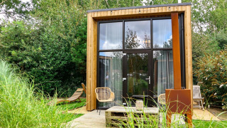Moderne tiny house med store glasvinduer, træfacade og udendørs siddepladser omgivet af grøn natur.