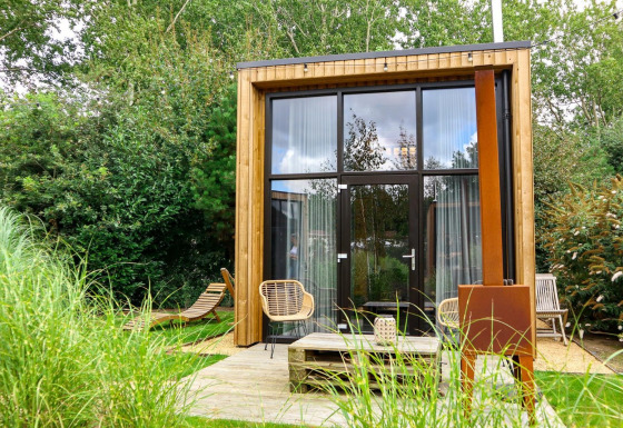 Tiny house moderna con grandi finestre in vetro, rivestimento in legno e area esterna circondata dal verde.