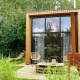 Moderne tiny house med store glasvinduer, træfacade og udendørs siddepladser omgivet af grøn natur.