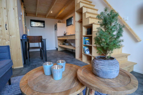 Wohnbereich im Tiny House bei Buitenhuizen, Niederlande, mit Holzmöbeln, Pflanze und Treppe.
