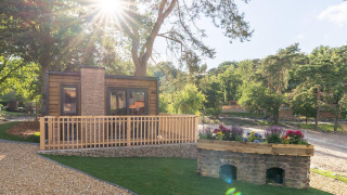 Zonnige dag bij Heidelodge tiny house, met bomen, grindpad en fleurige bloemen nabij het huisje.