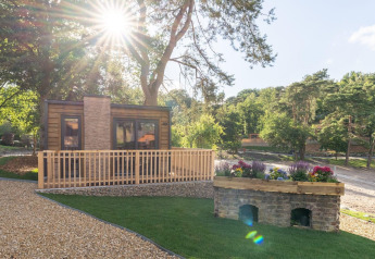 Día soleado en la tiny house Heidelodge, rodeada de árboles, camino de grava y flores coloridas cerca.