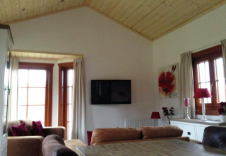 Woonkamer met houten plafond, grote ramen, tv aan de muur en rode accenten in Boekhorst Lodge, Brunssummerheide.