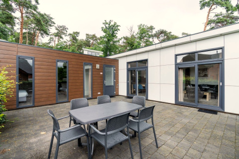 Buitenterras bij L-Pavilion op Brunssummerheide, Nederland, met eettafel, stoelen en uitzicht op een moderne lodge.