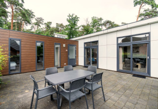 Buitenterras bij L-Pavilion op Brunssummerheide, Nederland, met eettafel, stoelen en uitzicht op een moderne lodge.