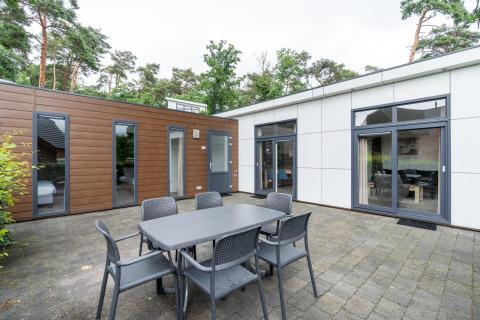Buitenterras aan L-Pavilion te Brunssummerheide, Nederland, met eettafel, stoelen en zicht op een moderne lodge.