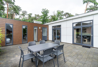 Buitenterras aan L-Pavilion te Brunssummerheide, Nederland, met eettafel, stoelen en zicht op een moderne lodge.