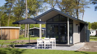 Modern tiny house met glazen voorgevel, terras, parasol en vier witte stoelen in groene natuur.