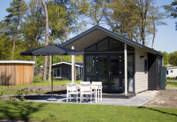 Tiny house moderna con grandi vetrate, patio esterno, ombrellone e quattro sedie bianche nel verde.