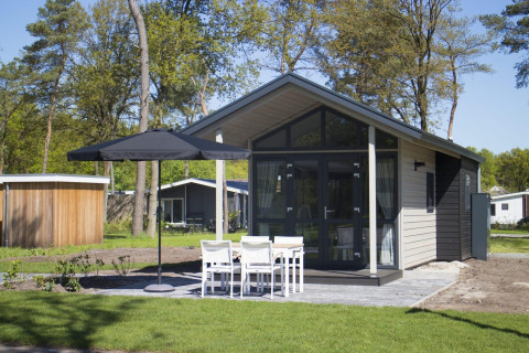 Modern tiny house met grote glazen gevel, terras, parasol en vier witte stoelen in een groene omgeving.