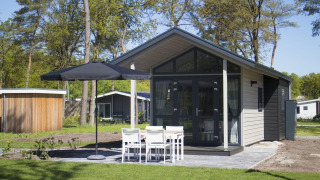 Modern tiny house met glazen voorgevel, terras, parasol en vier witte stoelen in groene natuur.