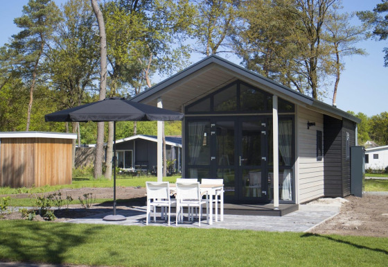 Tiny house moderna con grandi vetrate, patio esterno, ombrellone e quattro sedie bianche nel verde.