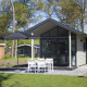 Moderne tiny house med stor glasfacade, terrasse, parasol og fire hvide stole, omgivet af natur.