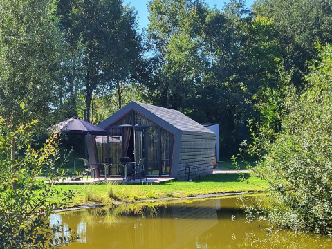 De Solo Retreat tiny house aan een vijver, omgeven door groene bomen en natuur onder de zon.
