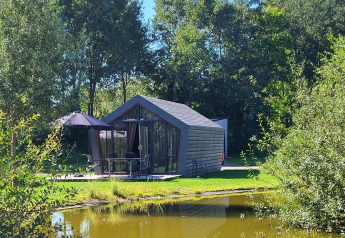 De Solo Retreat tiny house aan een vijver, omgeven door groene bomen en natuur onder de zon.