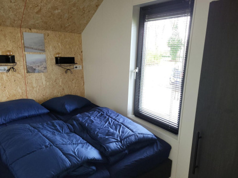 Piccola camera da letto con biancheria blu e grande finestra nella tiny house Solo Retreat a Reestervallei, Paesi Bassi.
