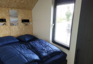 Piccola camera da letto con biancheria blu e grande finestra nella tiny house Solo Retreat a Reestervallei, Paesi Bassi.